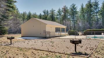 A small, single-story building with a flat roof, surrounded by a fenced area with a pool and dense pine trees in the background.