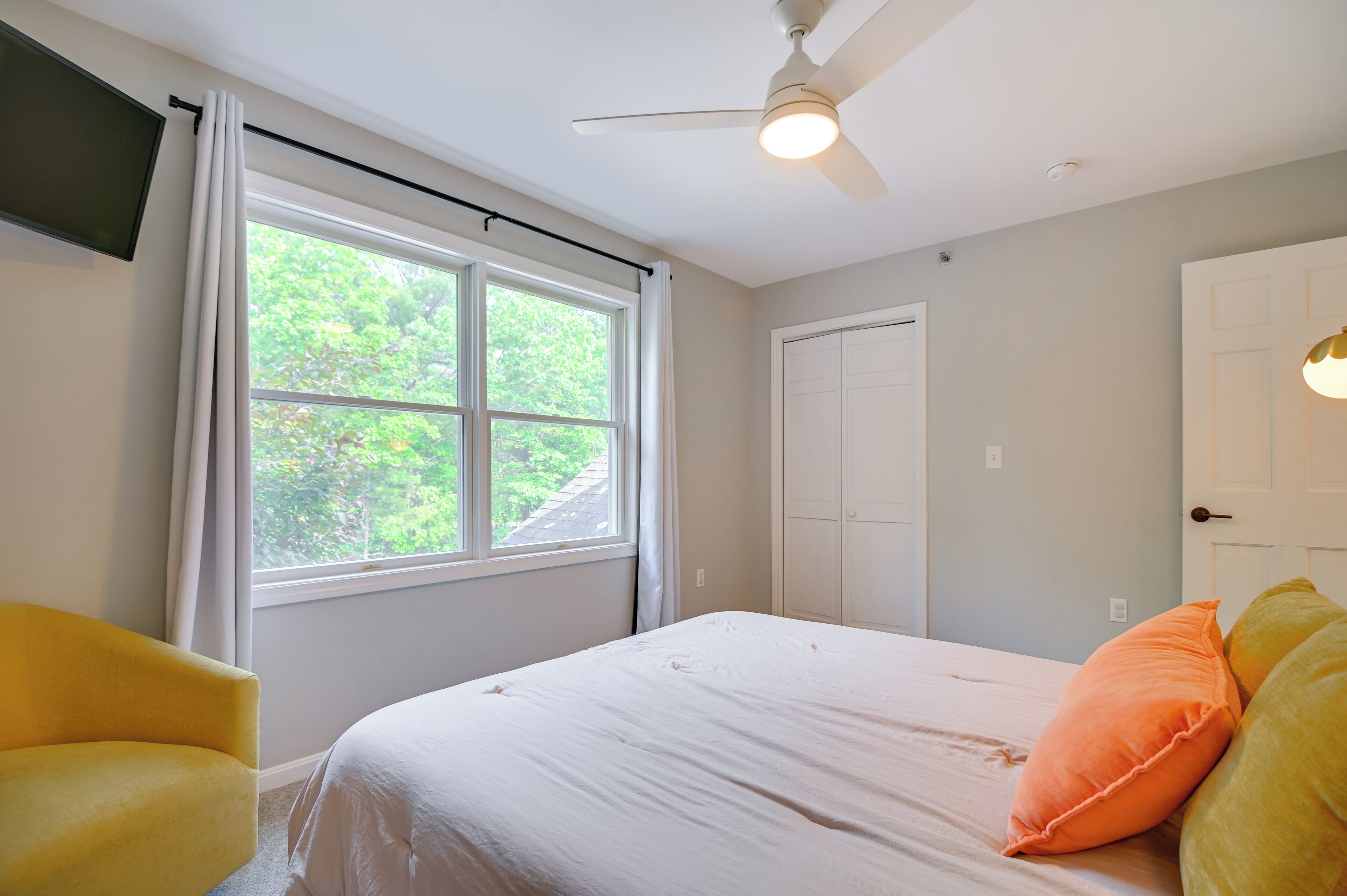 A bedroom with a bed, a ceiling fan, a window with curtains, a white door, and a yellow chair.