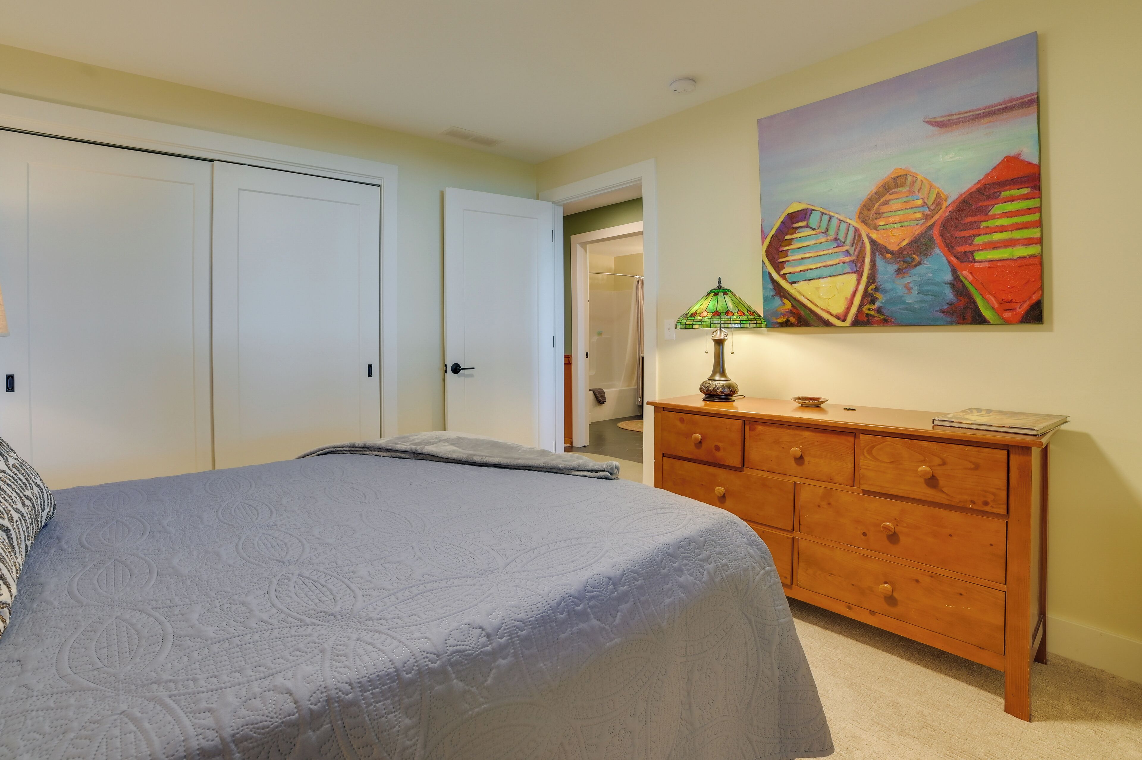 Un dormitorio con cama, cómoda, un cuadro y puerta que da a un baño.