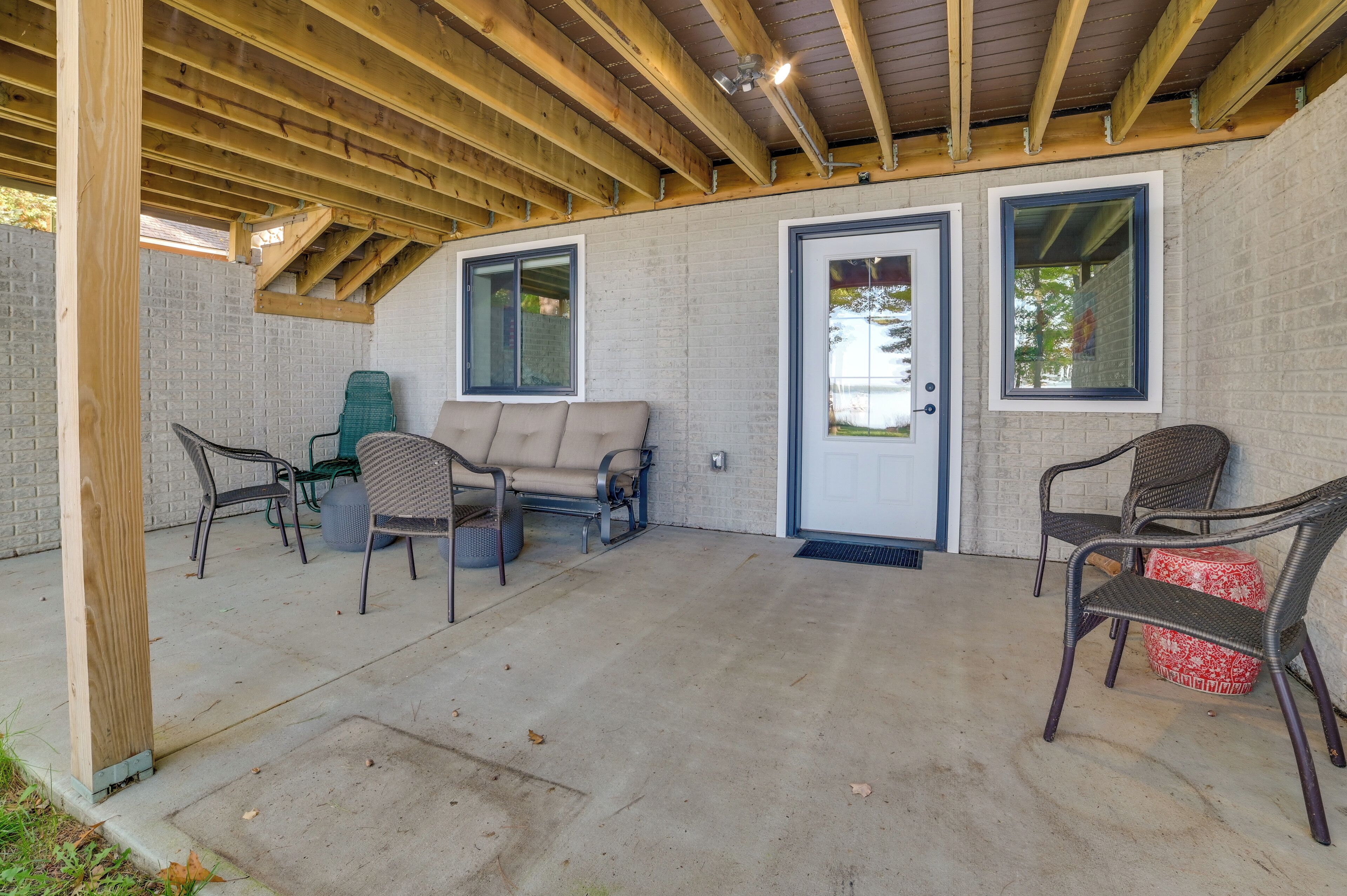 Un patio cubierto con piso de concreto, vigas de madera y pared de ladrillo. Se observan dos puertas, un banco y varias sillas.