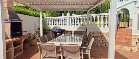 An outdoor patio with a pergola, a barbecue grill, and a dining table with chairs.