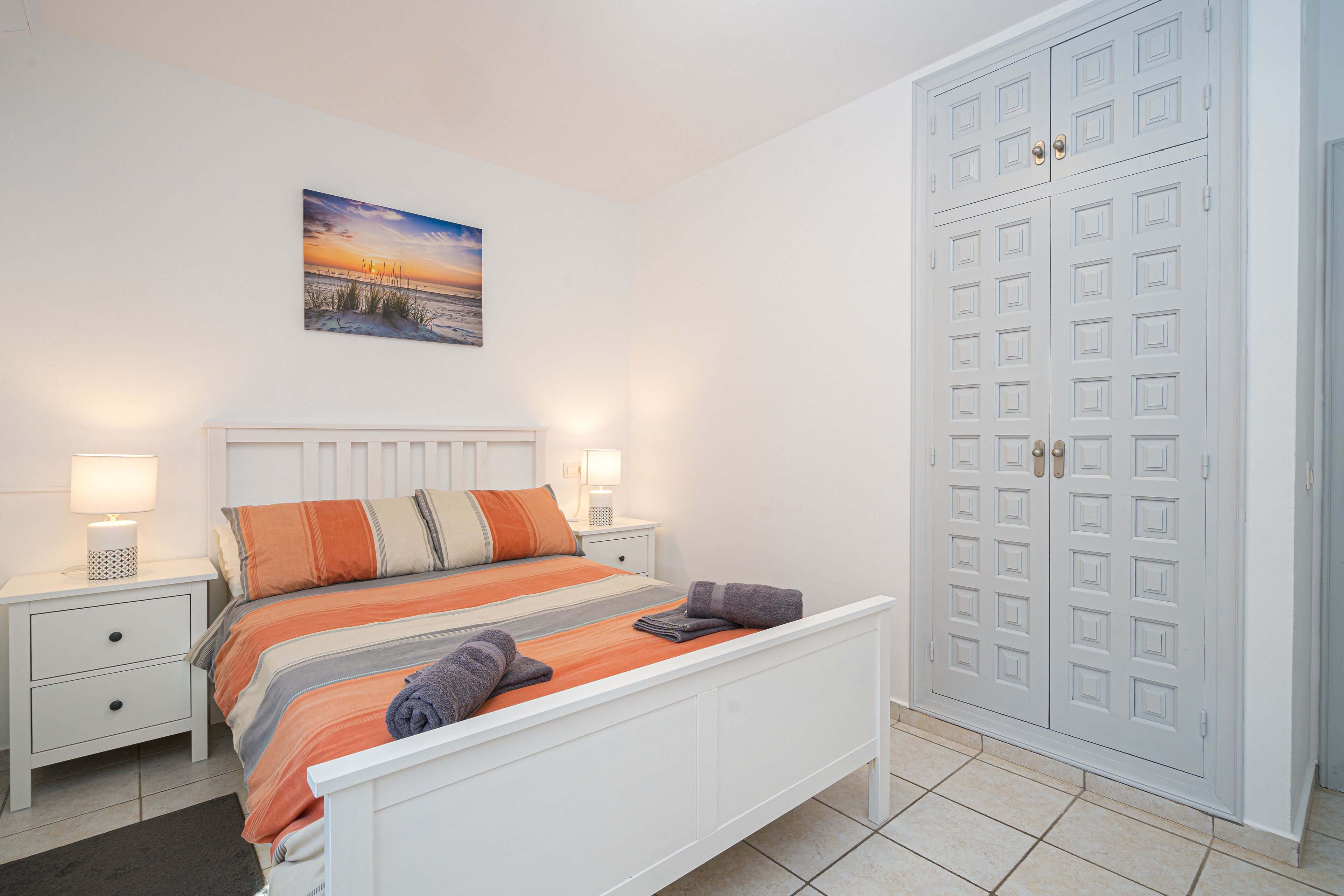 A neatly arranged bedroom with a bed, bedside tables, a painting, and a wardrobe.
