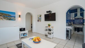 A modern living room with a TV, a dining table with fruit, a kitchen area with blue tiles, and a view of the sea.
