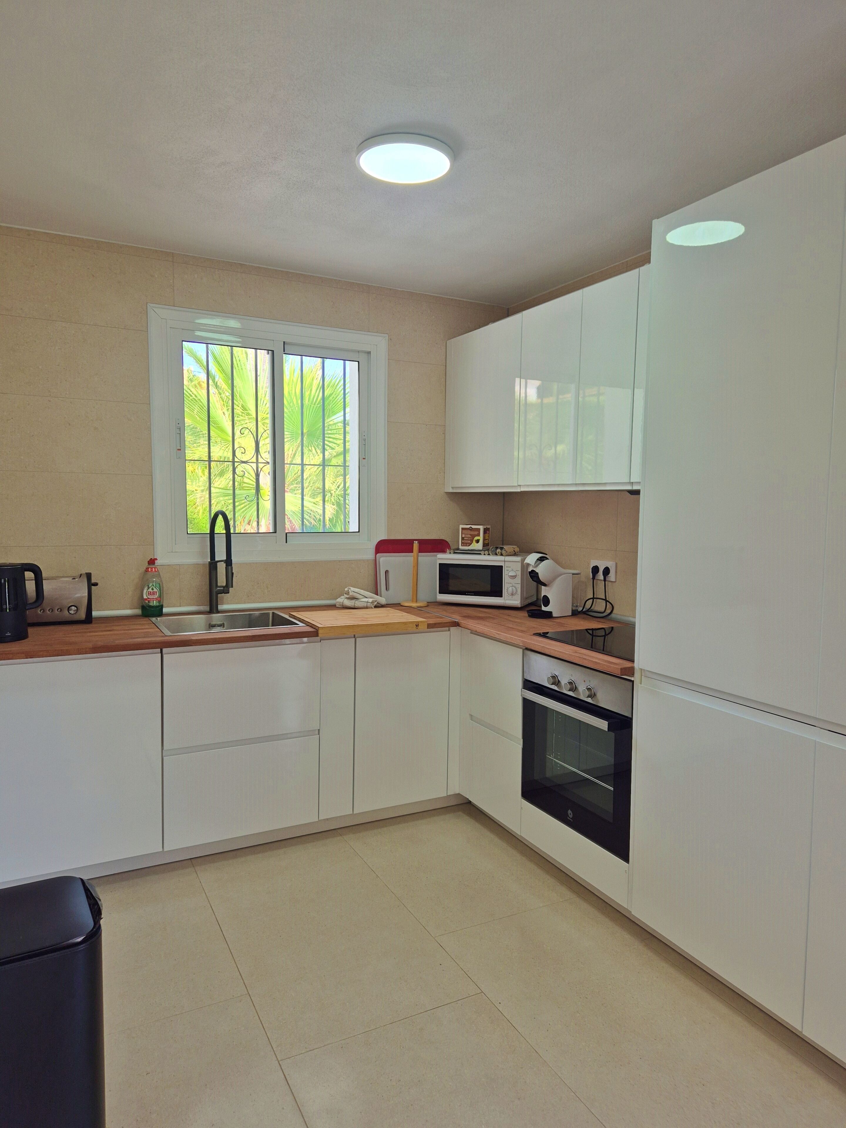 A kitchen with white cabinets, a microwave, an oven, a sink, and a window with a view of palm trees.