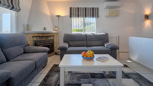 A living room with a fireplace, two grey sofas, a white coffee table with a fruit bowl, and a window with a striped valance.