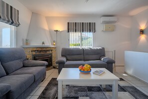 A living room with a fireplace, two grey sofas, a white coffee table with a fruit bowl, and a window with a striped valance. - Schöne Villa für 10 pax in der Nähe von Moraira mit Privatem Pool und Meerblick (Teulada)