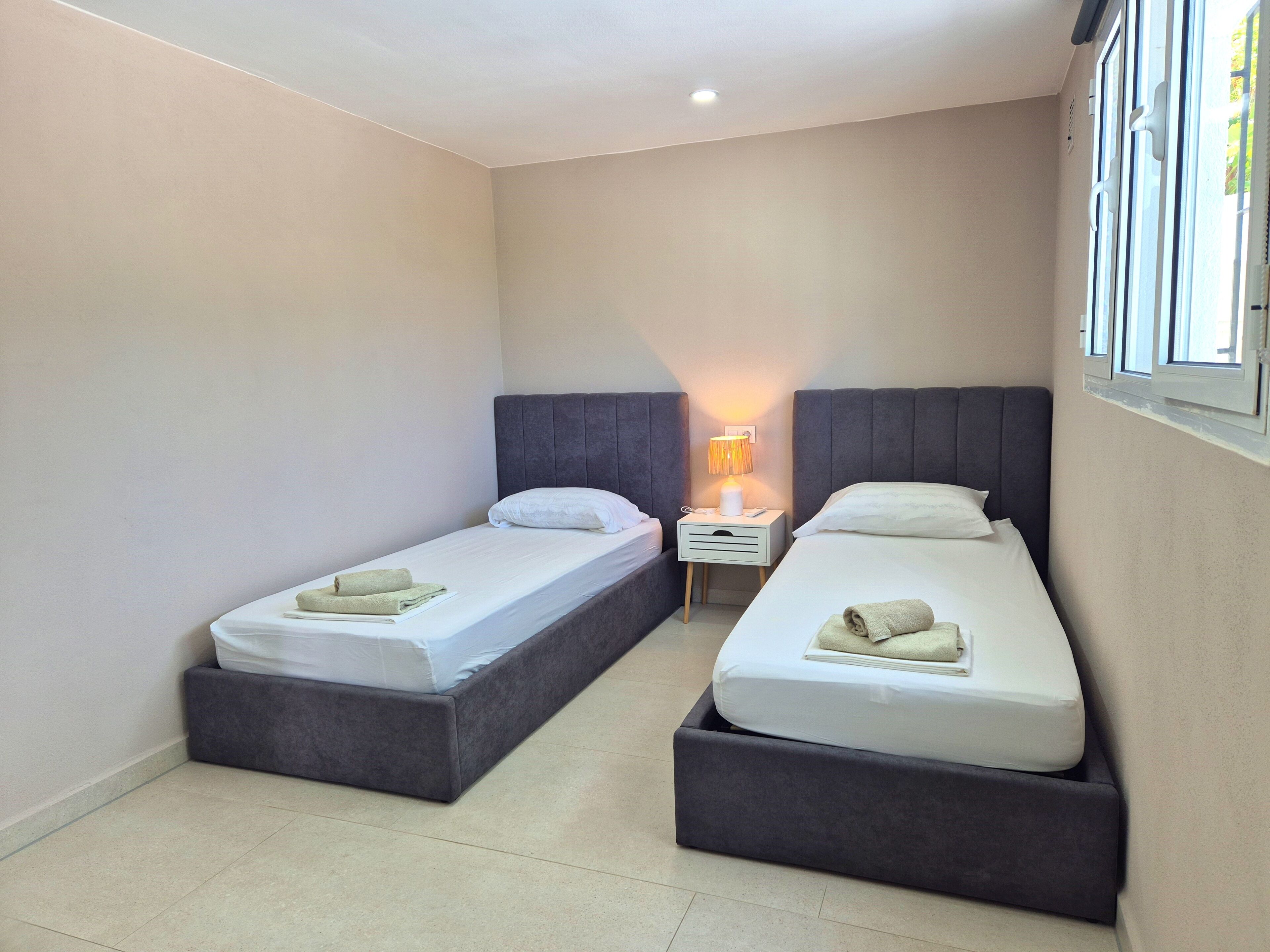 A small, modern bedroom with two single beds, a bedside table with a lamp, and a window with blinds.