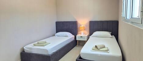 A small, modern bedroom with two single beds, a bedside table with a lamp, and a window with blinds.