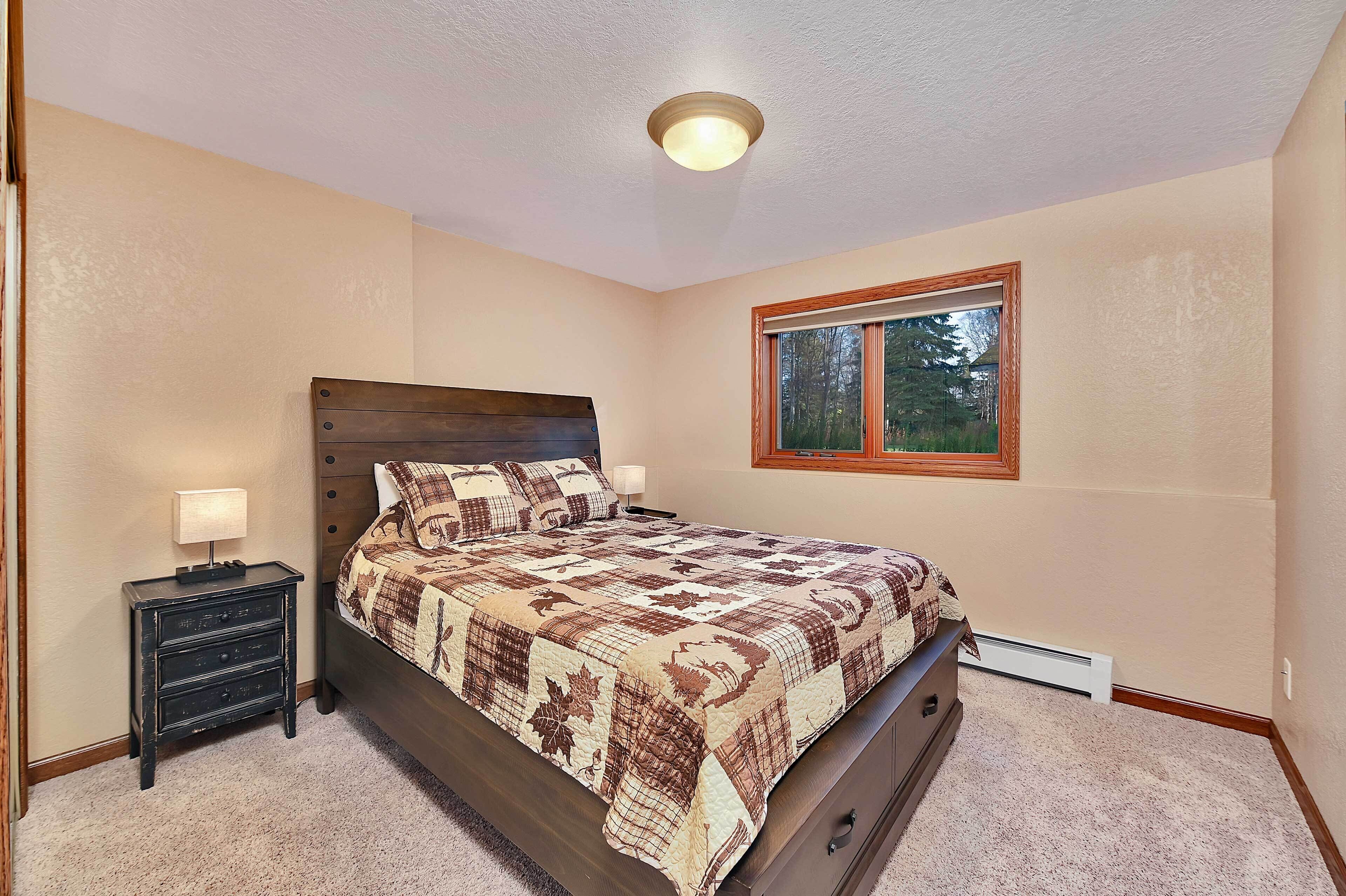A bedroom with a wooden bed, a nightstand with a lamp, and a window with a view of trees.