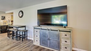 A living room with a flat-screen TV mounted on a cabinet, a dining area with a table and bar stools, and a clock on the wall.