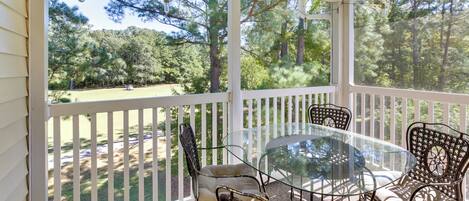 A covered patio with a glass table and wicker chairs, overlooking a grassy area with trees.