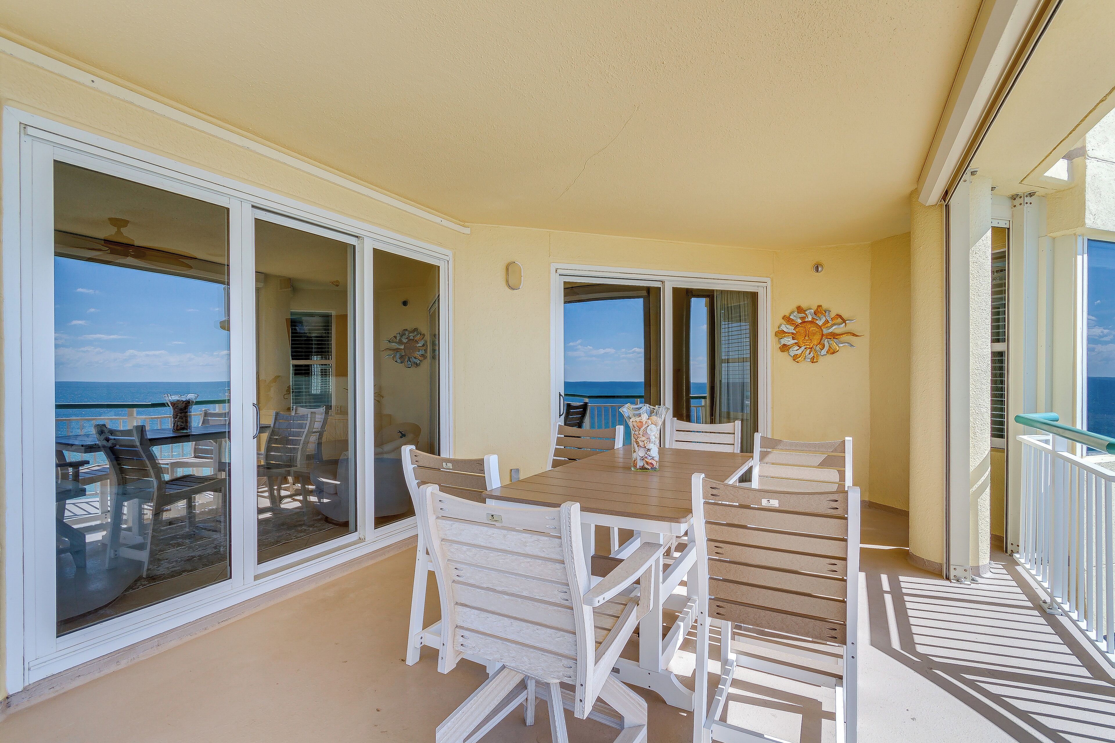 A balcony with a wooden table and chairs, a view of the ocean, and a decorative wall piece.