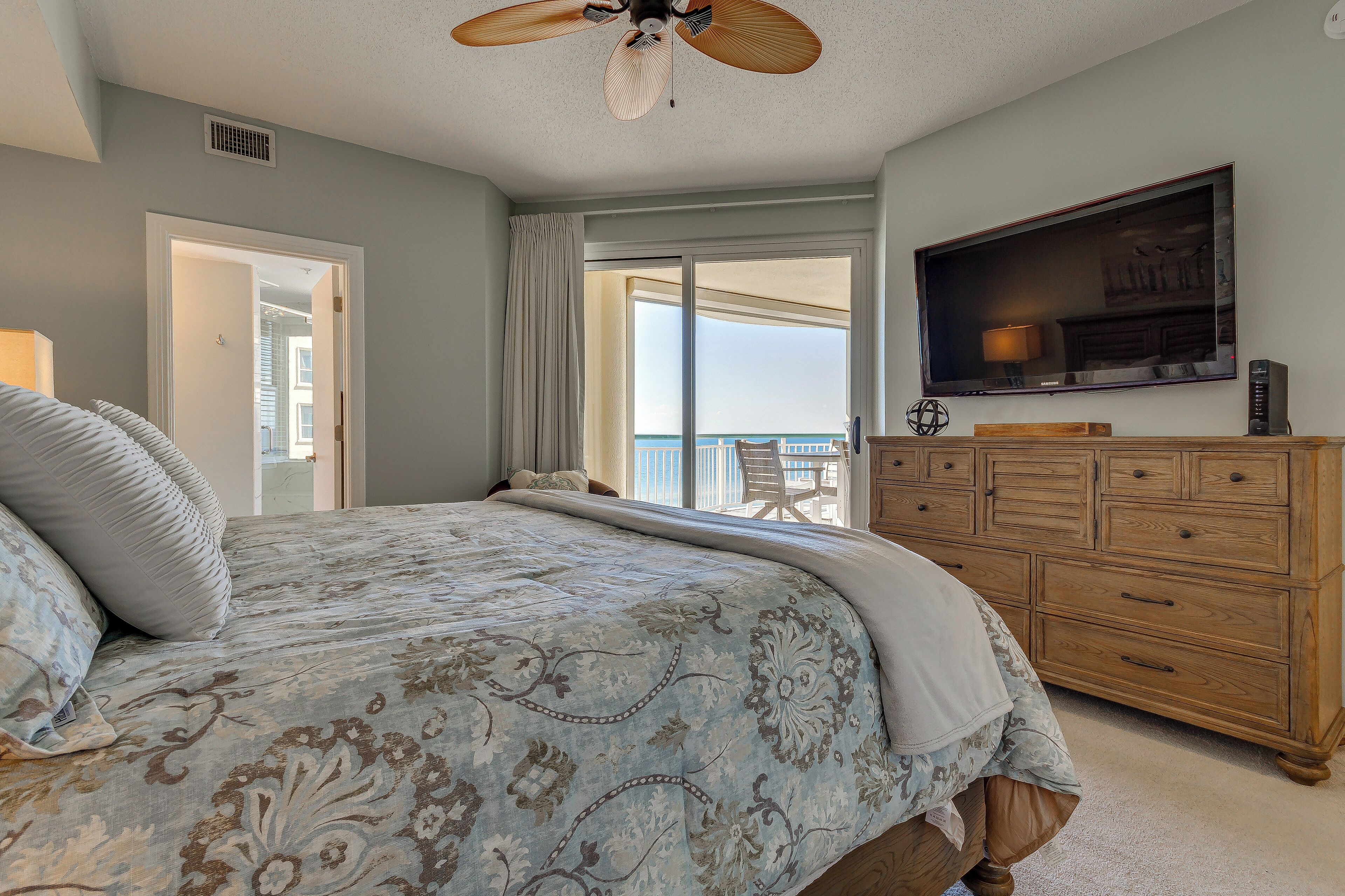 A bedroom with a large bed, a wooden dresser, a flat-screen TV, and a balcony with a view.