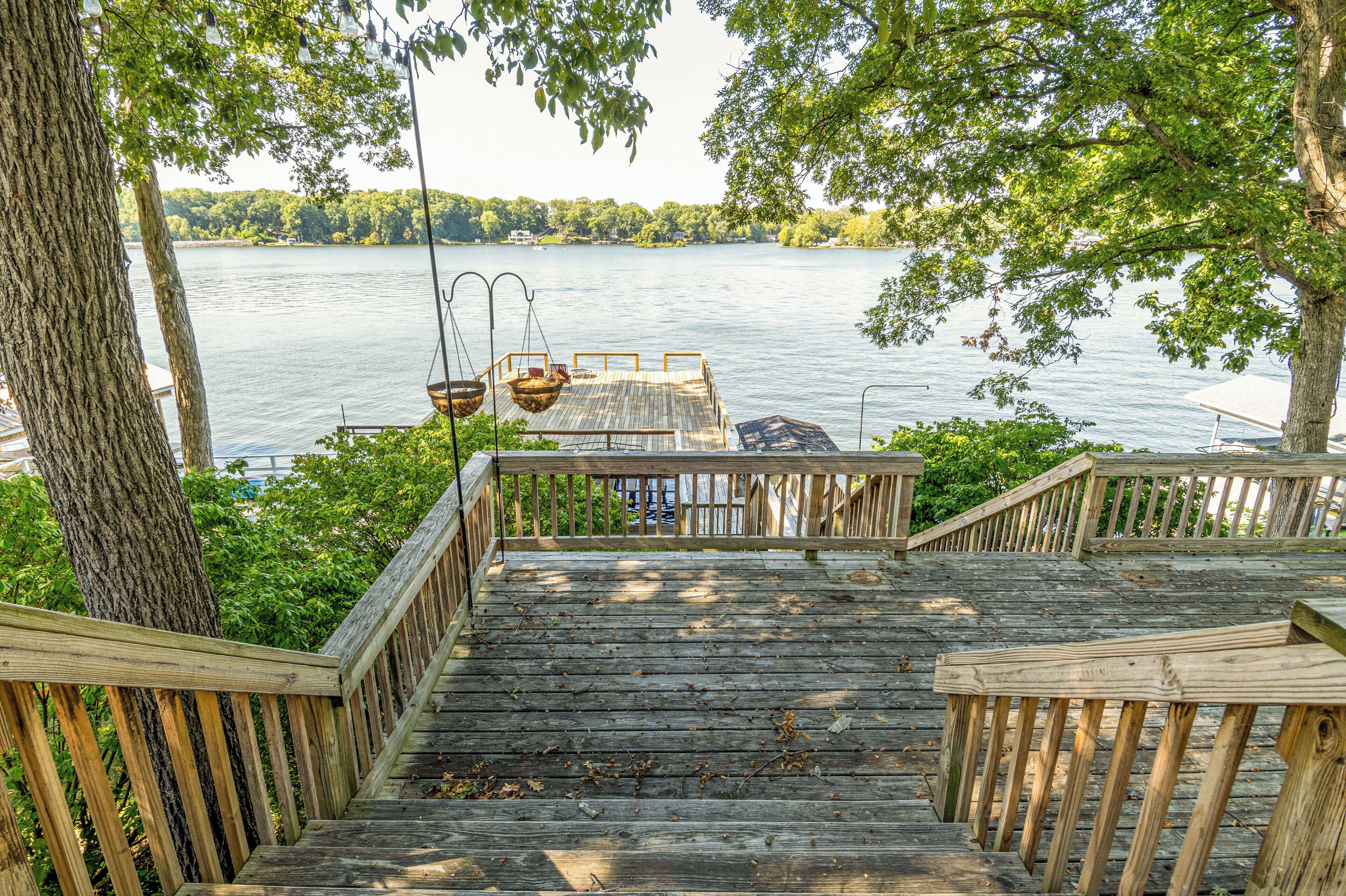 A wooden deck with a view of a lake, a hanging chair, and a table.