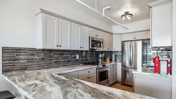A modern kitchen with white cabinets, a granite countertop, stainless steel appliances, and a backsplash with a wood-like pattern. - Mountain-view Deck: Stylish Virginia City Apt! (Virginia City)