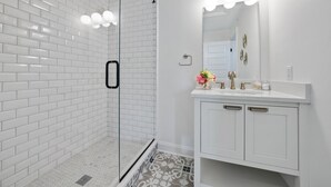 A modern bathroom with white subway tiles, a glass shower enclosure, a white vanity with gold fixtures, and a mirror with three lights. - Stylish Boutique Style Condo (Sandusky)