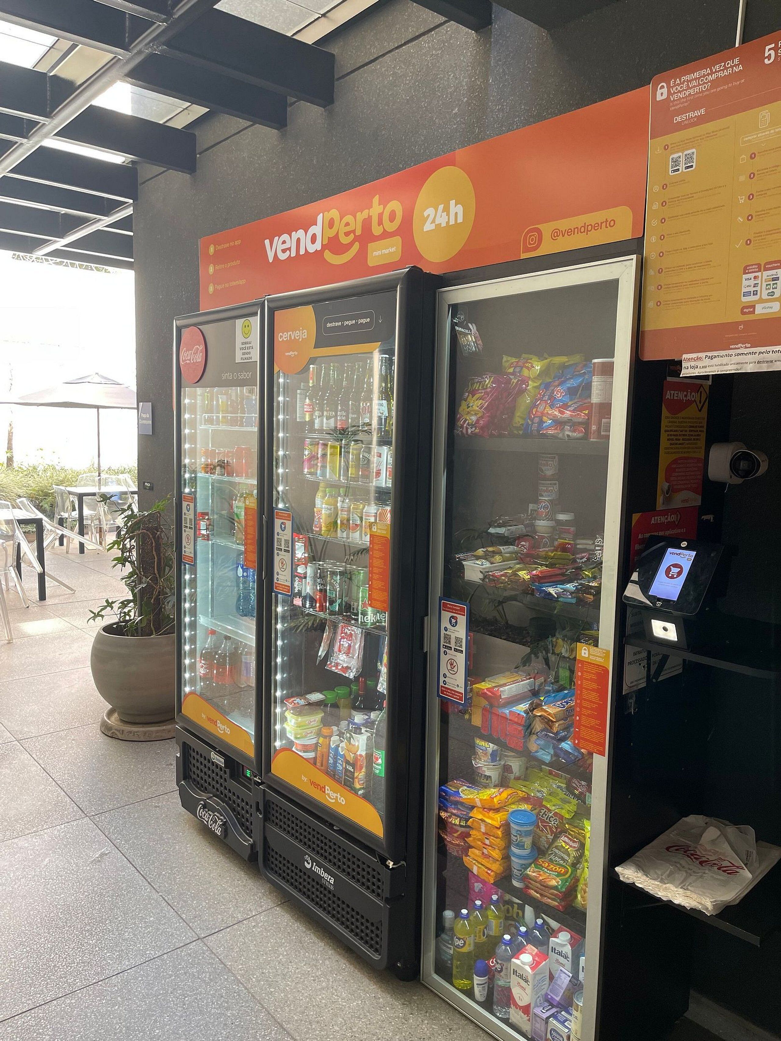 Máquina de vendas com o logotipo da Coca-Cola, identificada com 'vend perto' e '24h', indicando um serviço próximo e disponível 24 horas.