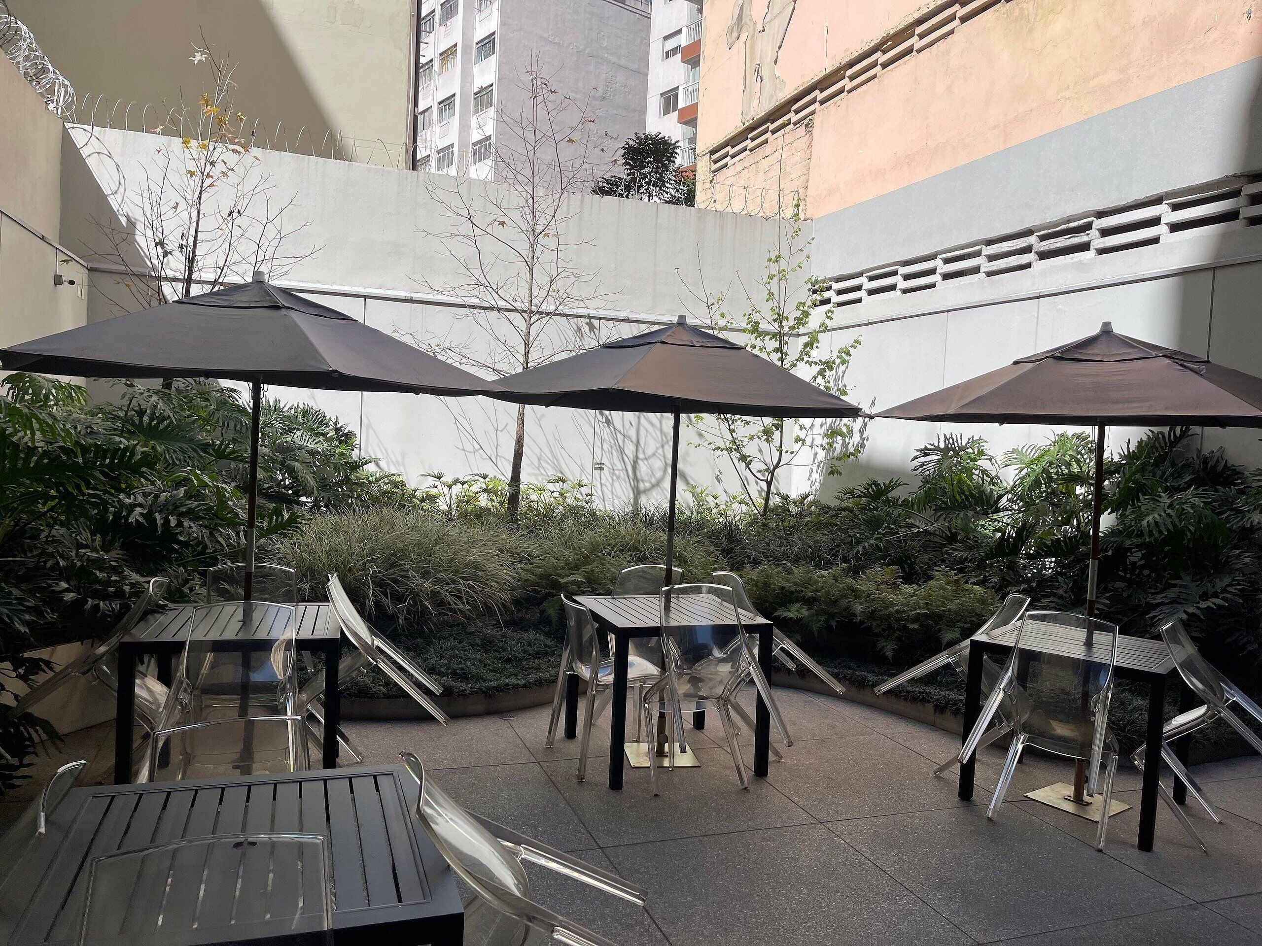 Un'area patio esterna con tavoli e sedie sotto ombrelloni, circondata da vegetazione e un edificio sullo sfondo.