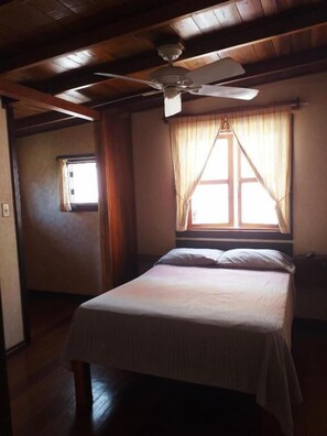Un lit simple dans une chambre avec des poutres apparentes, un ventilateur de plafond et une fenêtre avec des rideaux.