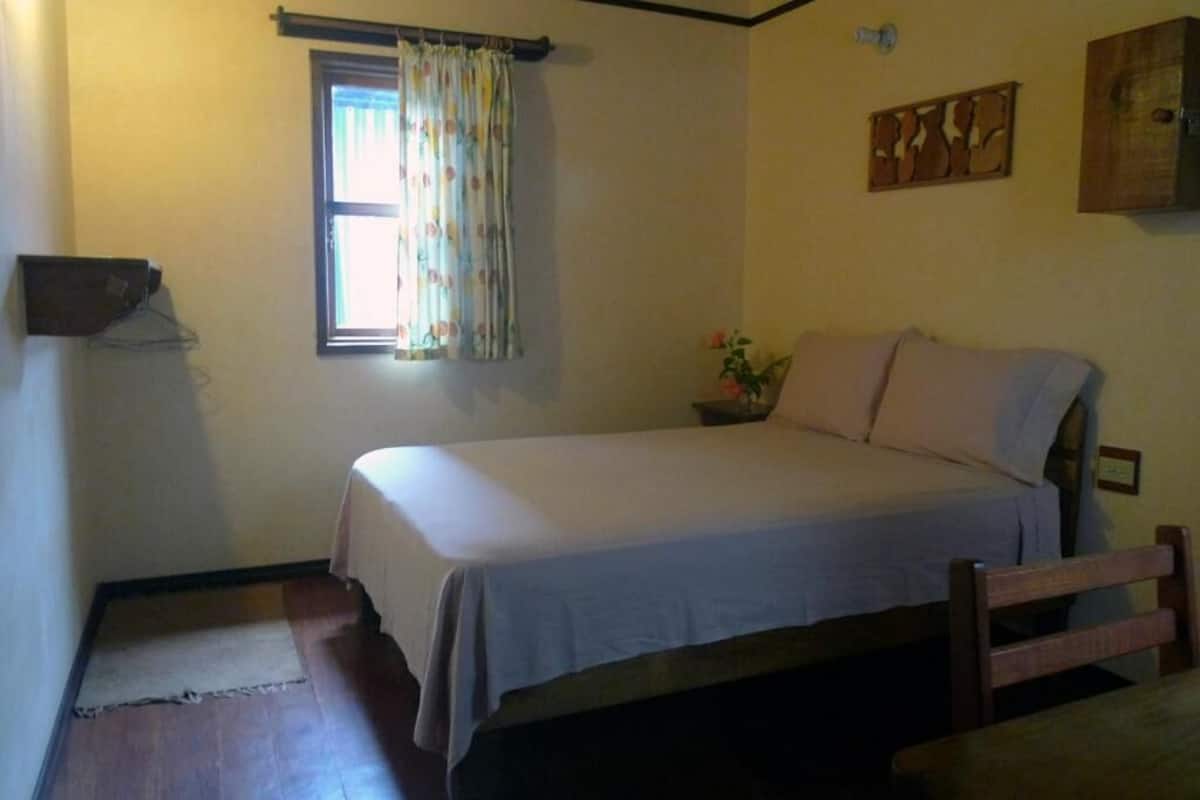 A bedroom with a single bed, a window with curtains, a bedside table, and a wall-mounted shelf.