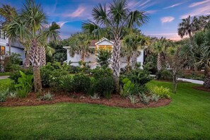 A well-maintained garden with palm trees and various shrubs, a white house with a yellow door, and a clear sky.