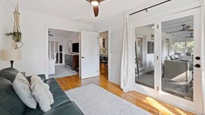 Un salón espacioso con un sofá verde oscuro, cojines blancos y un ventilador de techo. Un amplio ventanal corredera da a un área exterior.