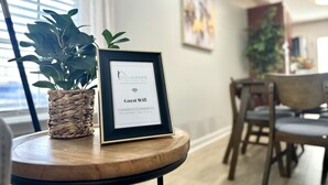 Señal enmarcada que dice 'Guest Wifi' sobre una mesa de madera con una planta en maceta a un lado, en una habitación con un comedor al fondo.