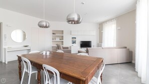 A modern living room with a wooden dining table, white chairs, a sofa, and a TV.