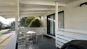 A covered outdoor area with a table and chairs, a black grill, and a view of the street and greenery.