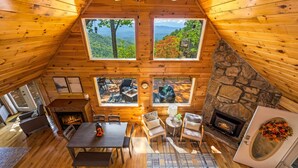 A cozy log cabin with a stone fireplace, wooden walls, and large windows offering scenic views. - Top of the Smoky Mountain Views, Hot Tub + 10ft Projector (Sevierville)