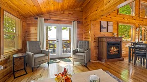 A cozy cabin interior with wooden walls, a fireplace, two armchairs, a dining table, and a large window overlooking a deck.
