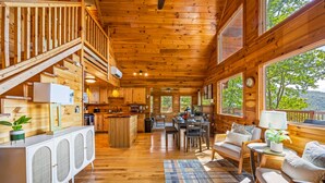 A spacious wooden cabin with a dining area, kitchen, and a large window overlooking greenery. - Top of the Smoky Mountain Views, Hot Tub + 10ft Projector (Sevierville)
