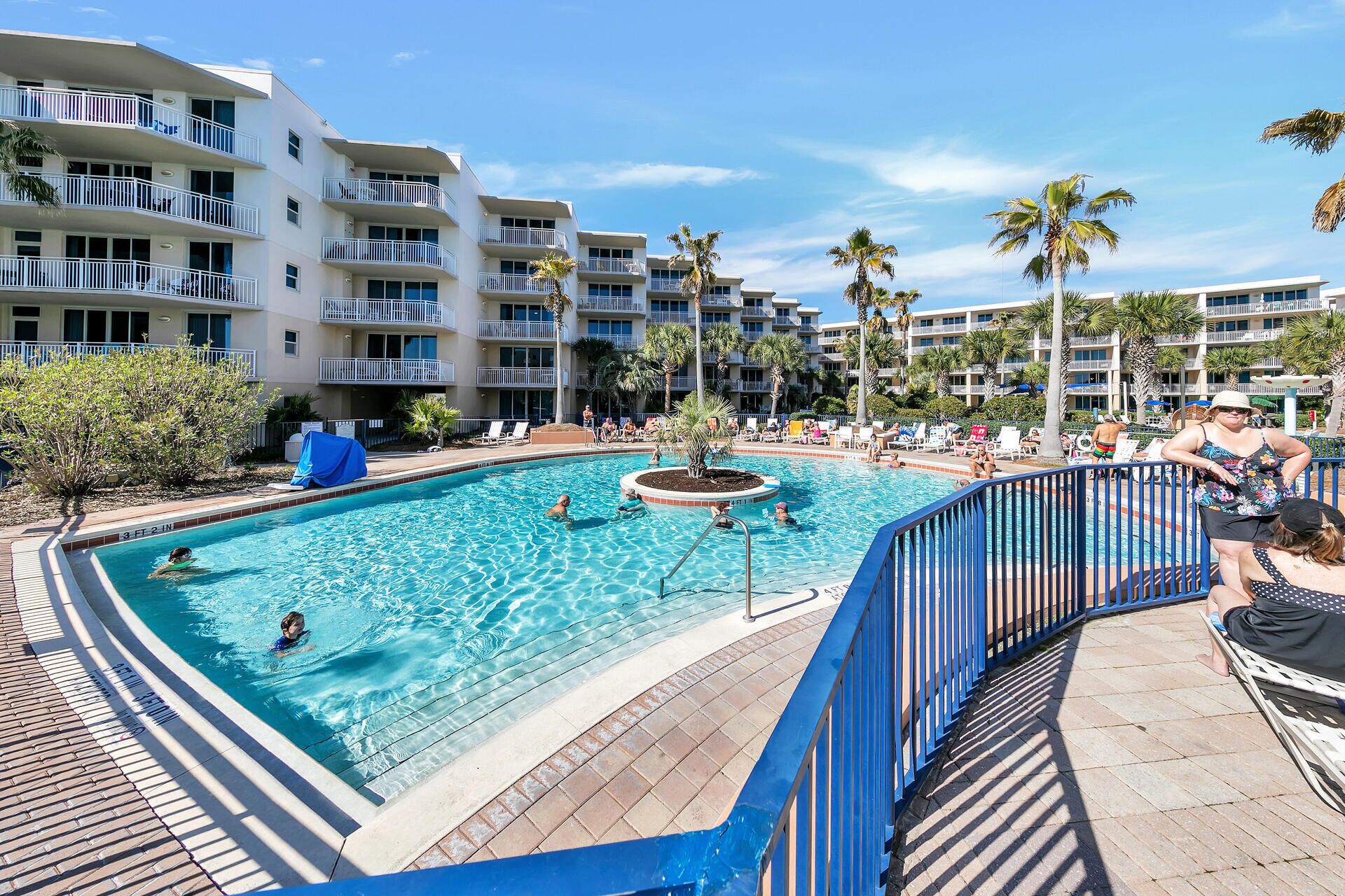Une piscine avec une terrasse bleue, entourée de palmiers et d’un immeuble à plusieurs étages doté de balcons.