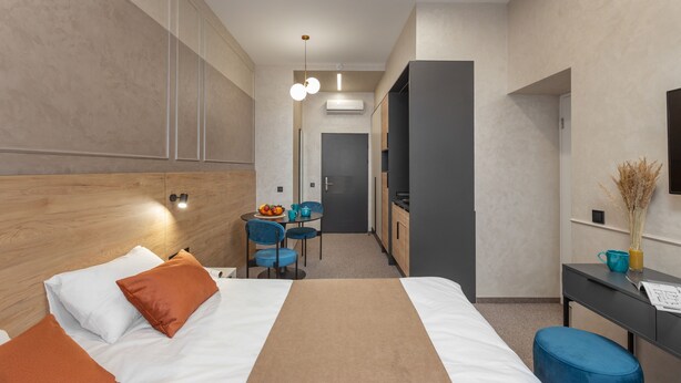 Habitación de hotel moderna con una cama grande, un escritorio con una silla, una mesita con frutero y una kitchenette.