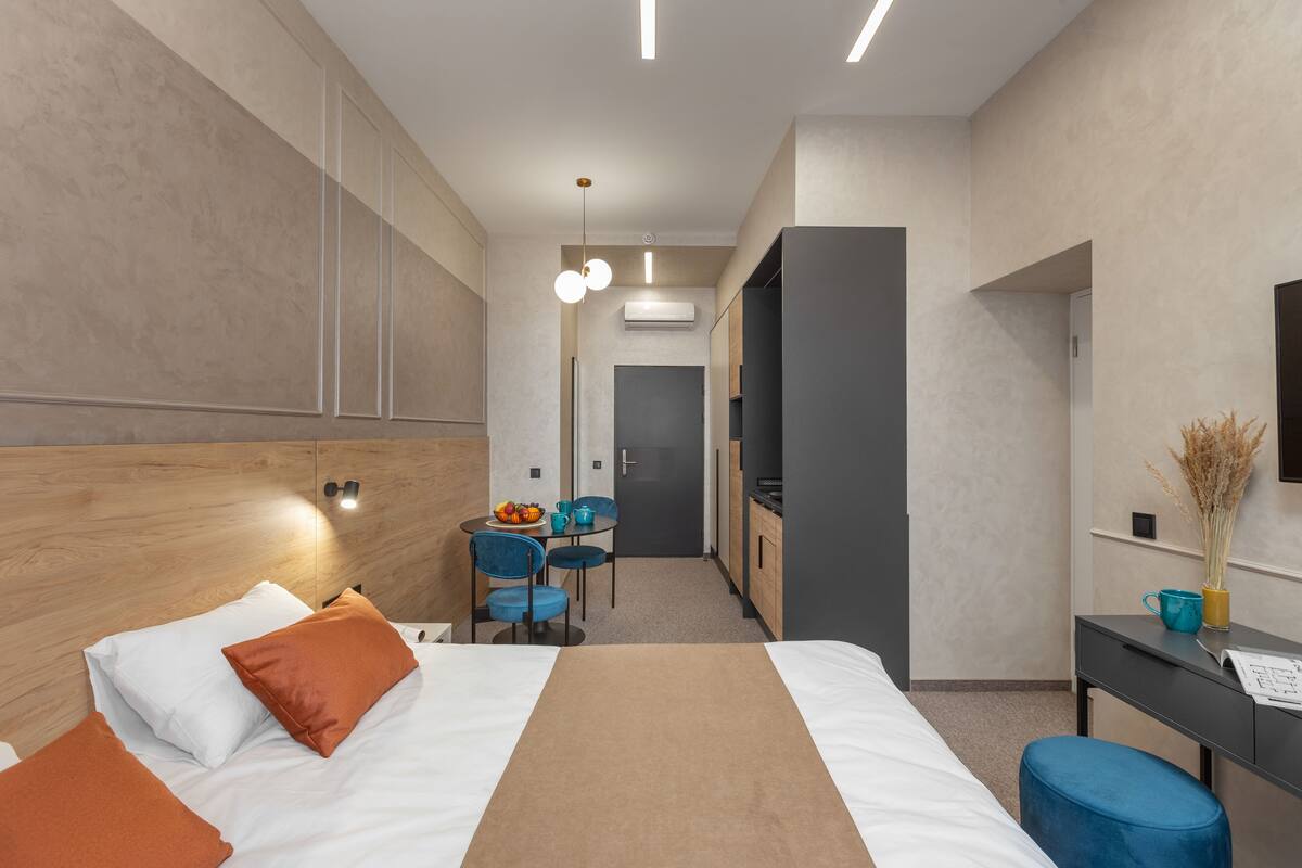 A modern hotel room with a large bed, a desk with a chair, a small table with a fruit bowl, and a kitchenette area.