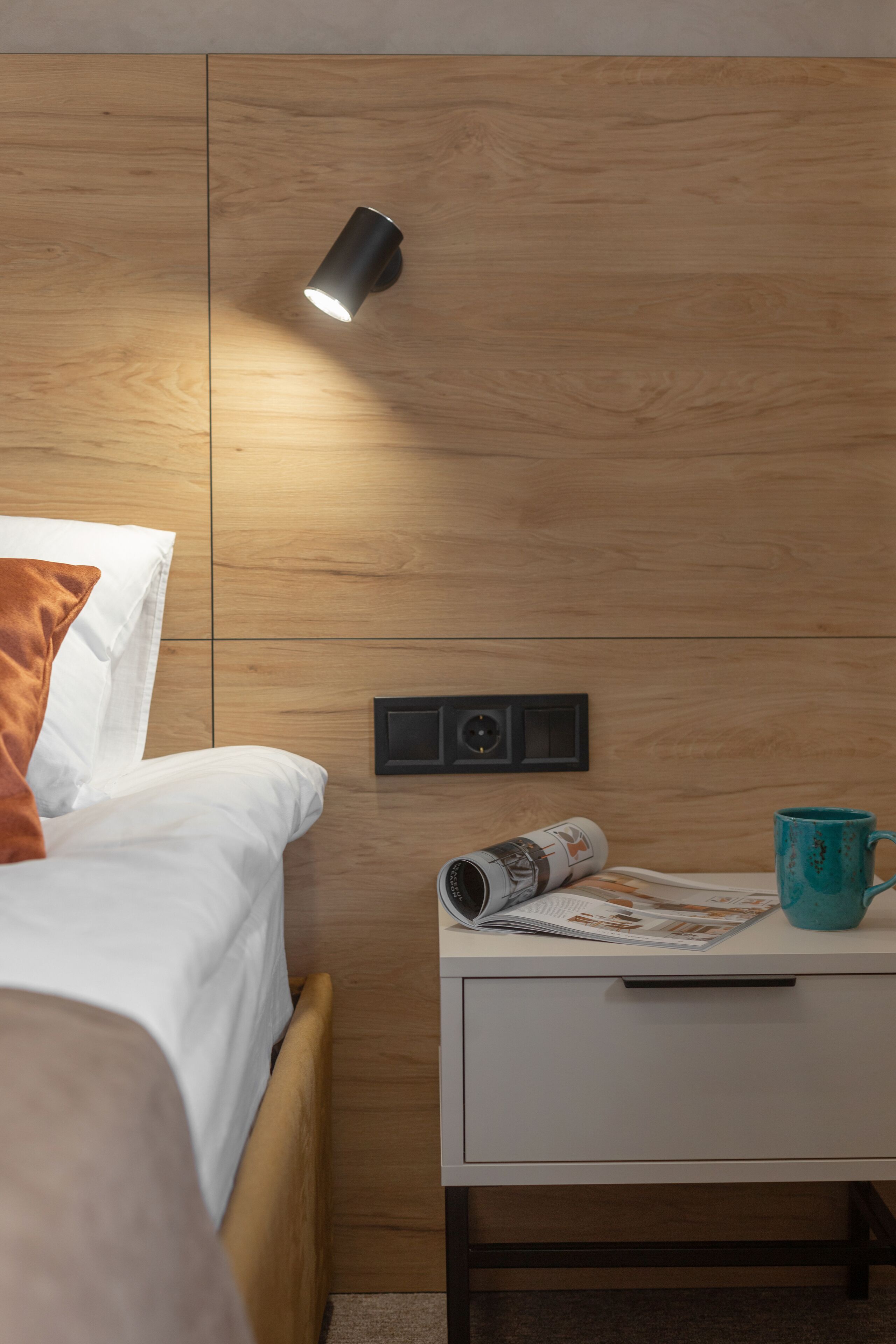 A neatly made bed with white bedding and a brown pillow, a bedside table with a magazine and a teal mug, and a wall-mounted light fixture.