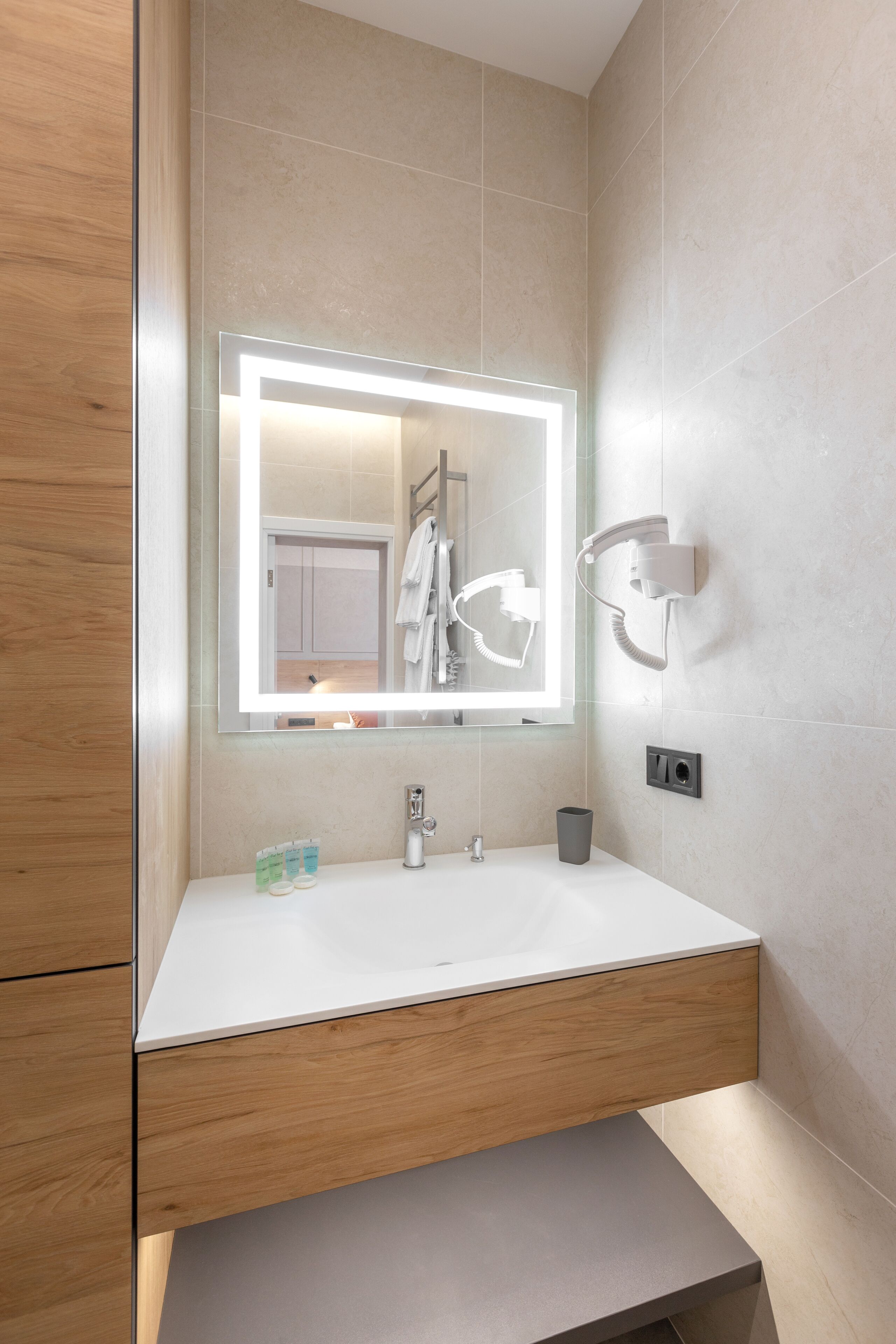 A modern bathroom with a white sink, wooden vanity, a mirror with integrated lighting, a towel rack, and a hairdryer mounted on the wall.