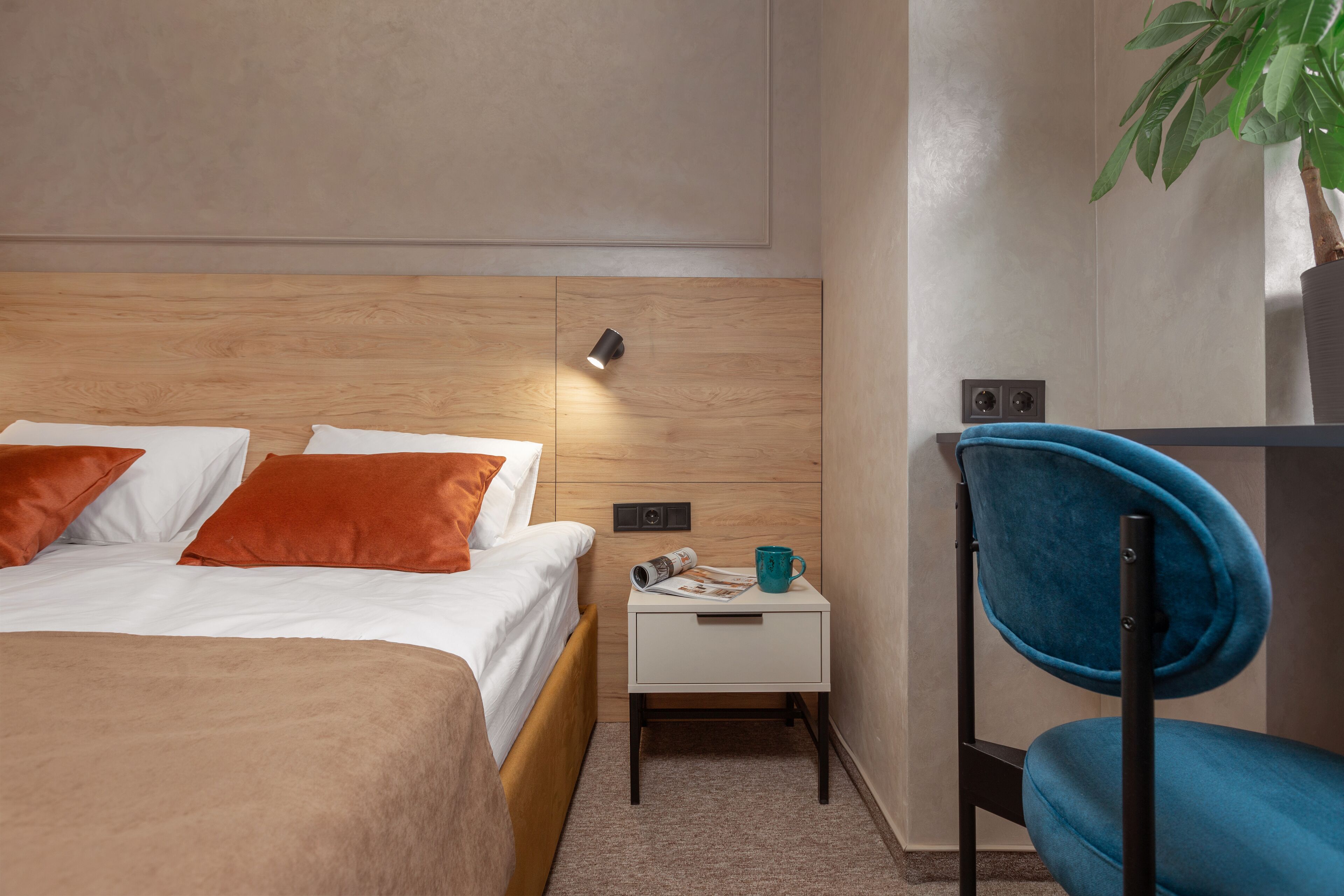 Habitación de hotel ordenada con cama, mesita de noche pequeña, una silla y una lámpara fijada a la pared.