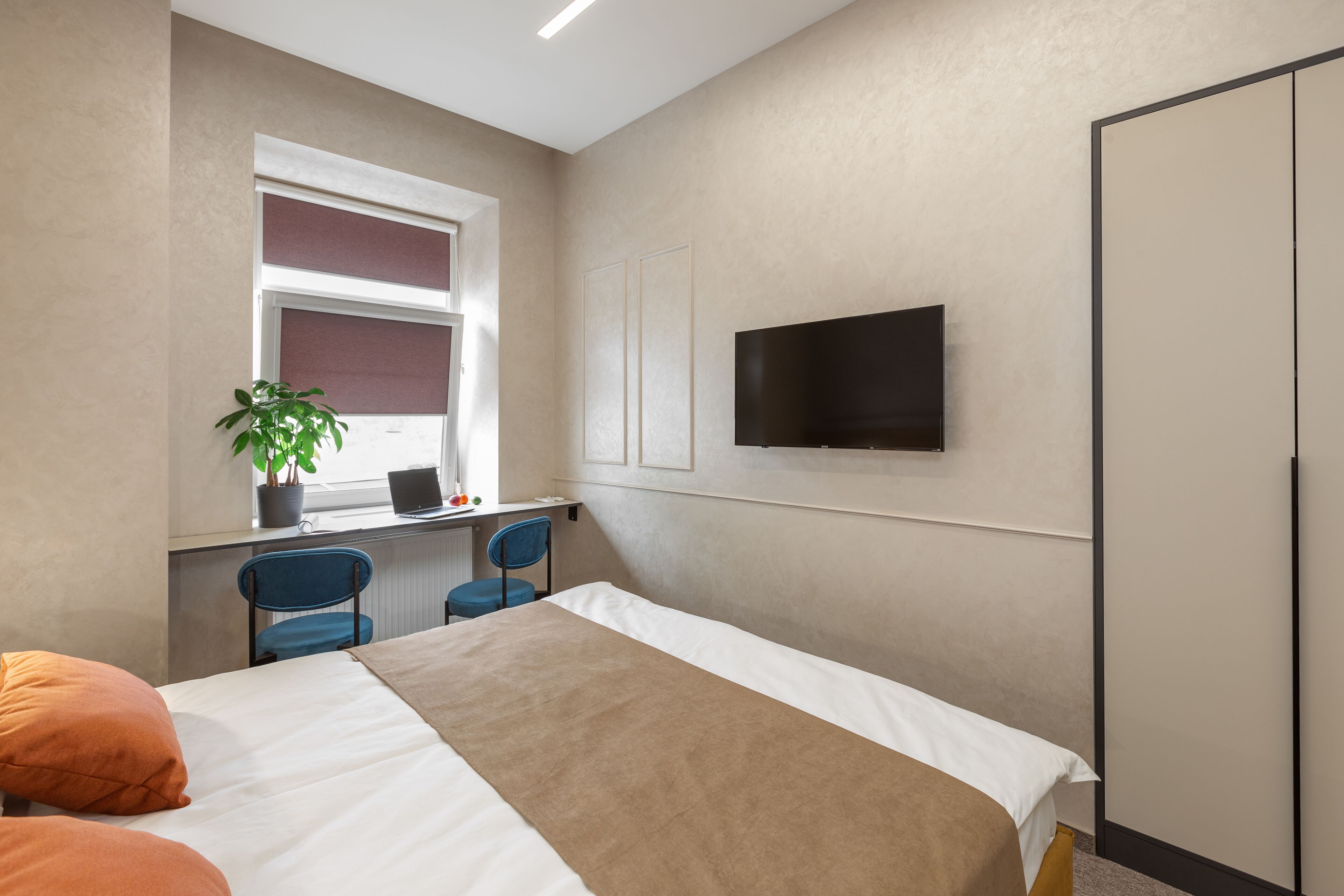 Habitación de hotel con una cama, un escritorio con computadora, dos sillas, un televisor y una ventana con persianas.