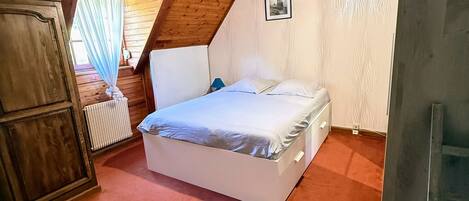 Une chambre avec un plafond en pente, un lit, un radiateur et une fenêtre avec des rideaux.