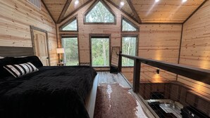 A bedroom with a bed, a nightstand, a lamp, a window, and a view of the outdoors. - The Beautiful Moonlight Dream Cabin in Broken Bow is STUNNING! (Broken Bow)