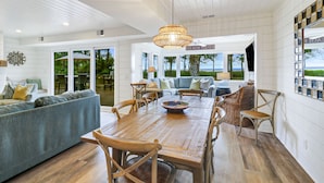 A spacious living room with a wooden dining table, comfortable seating, and large windows offering a view of the outdoors.