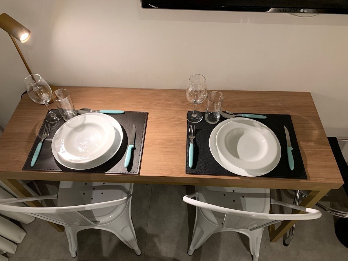 A dining table set for two with white chairs, black placemats, and teal-handled cutlery.