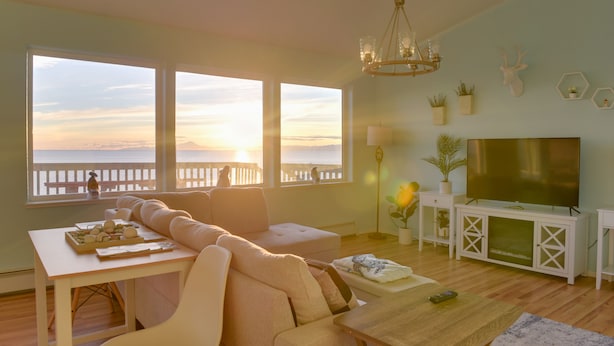 Een woonkamer met een bank, een televisie en een eettafel. Er zijn grote ramen met uitzicht op de zonsondergang.
