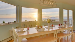 Een eetruimte met uitzicht op de ondergaande zon boven de oceaan, een houten tafel, witte stoelen en een hanglamp.