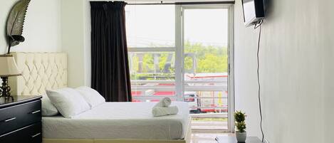 Habitación de hotel con dos camas, un televisor montado en la pared, una mesita con una planta y vista a un estacionamiento a través del ventanal.