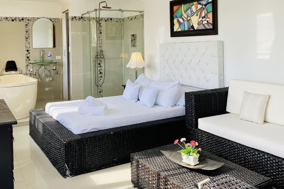 A modern hotel room with a large bed, a sofa, a small table with flowers, a shower area, and a bathroom with a bathtub.