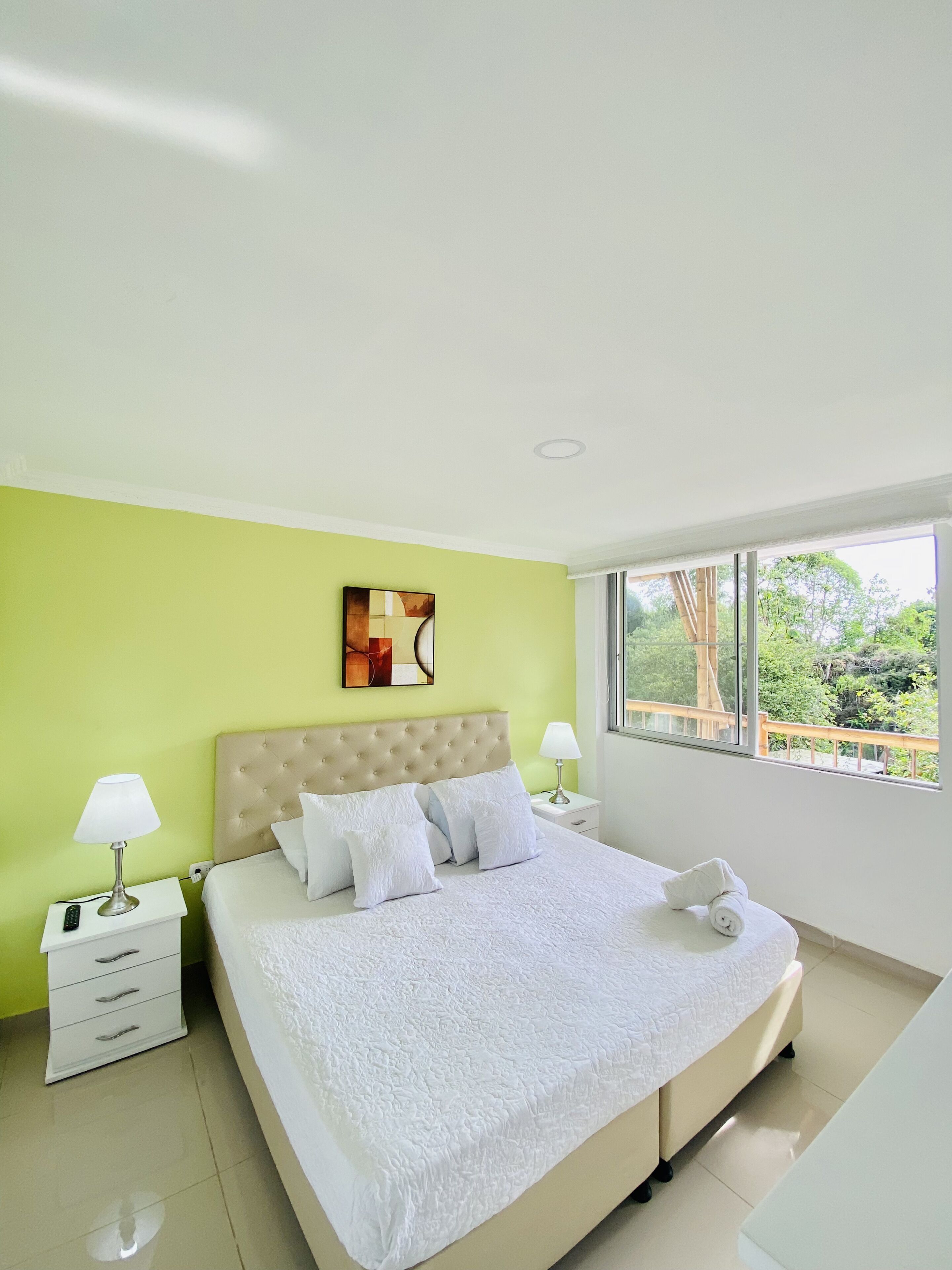 A modern bedroom with a large bed, a green accent wall, a window with a view, and a painting above the bed.