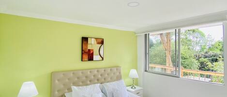A modern bedroom with a large bed, a green accent wall, a window with a view, and a painting above the bed.