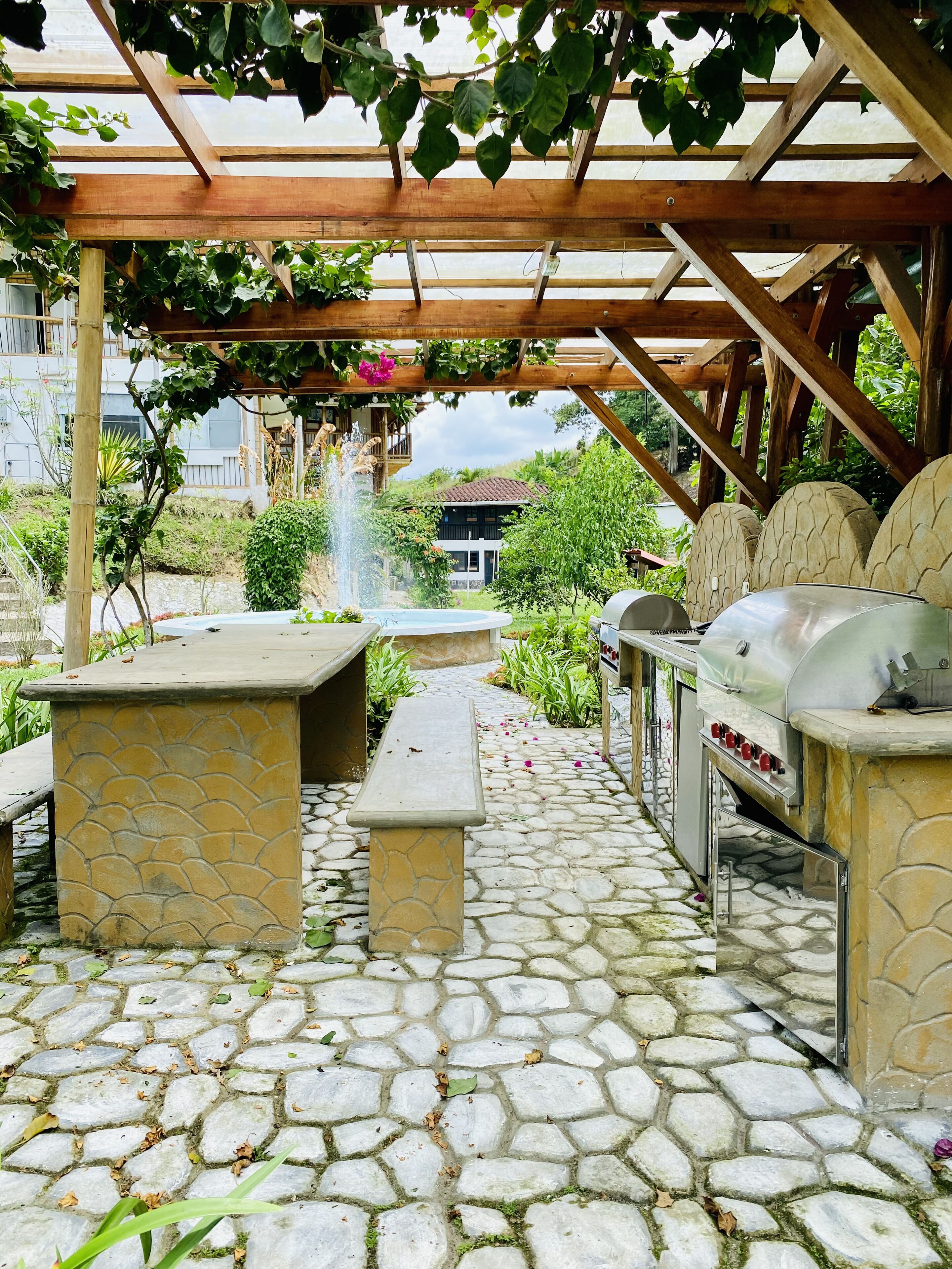An outdoor patio with stone flooring, a wooden pergola, a stone bench, and a barbecue grill.